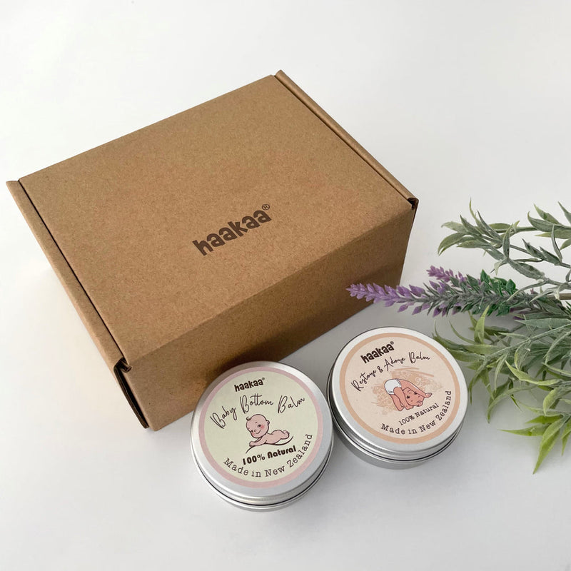 A box with two Haakaa baby skincare products in front of it - Baby Bottom Balm and Restore and Adore Balm. Beside them is a sprig of lavender.
