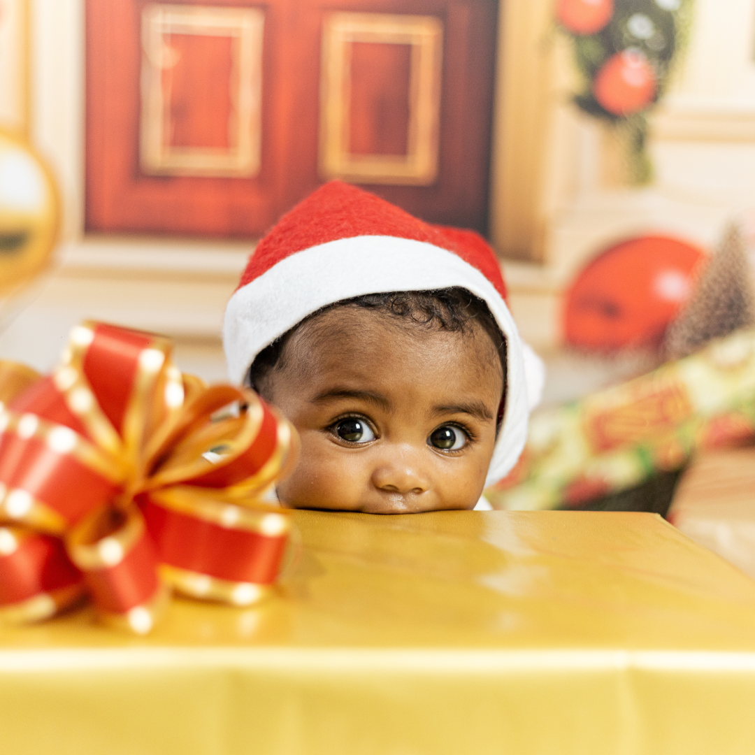 A cute baby wearing a Santa hat is biting on the edge of a gold paper-wrapped present